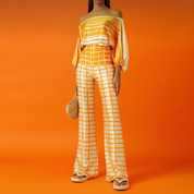 Golden-orange tie-dye coord set featuring a wide boat-neck blouse with voluminous sleeves and high-waisted wide-leg pants with grid print, styled with platform sandals and a beige woven handbag on a vibrant orange studio background.