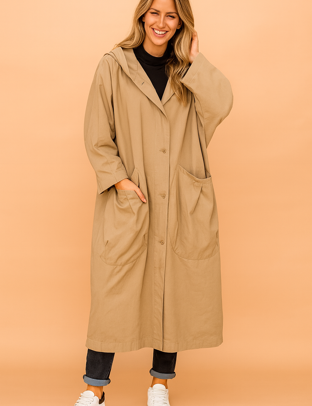 Oversized beige hooded trench coat with button closure, large front patch pockets, paired with a black turtleneck, cuffed dark denim jeans, and white sneakers.