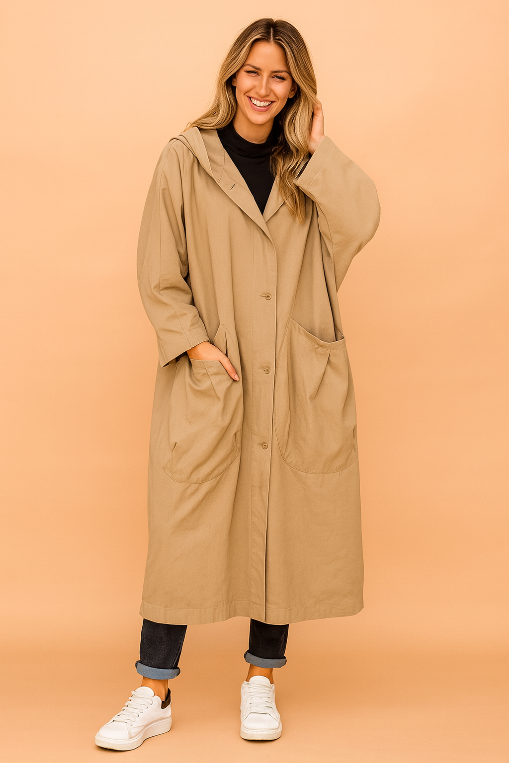Oversized beige hooded trench coat with button closure, large front patch pockets, paired with a black turtleneck, cuffed dark denim jeans, and white sneakers.