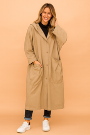 Oversized beige hooded trench coat with button closure, large front patch pockets, paired with a black turtleneck, cuffed dark denim jeans, and white sneakers.