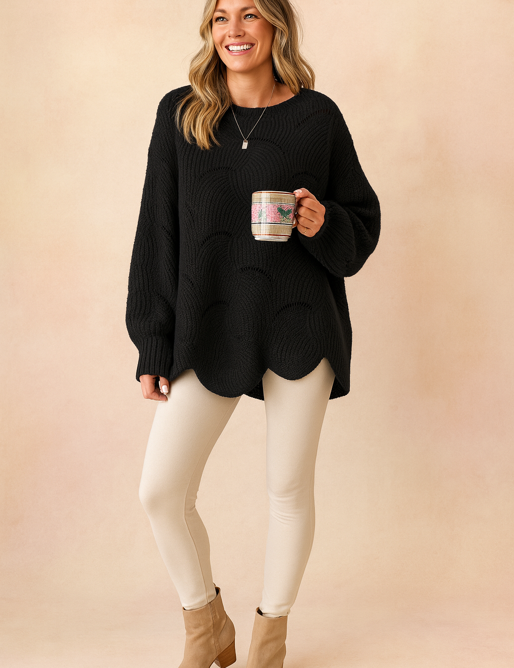 Oversized black knit sweater with circular hole pattern and scalloped hem, paired with cream-colored leggings and tan suede block-heel ankle boots.