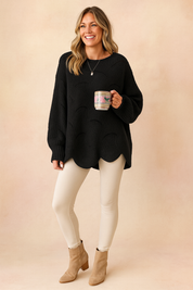 Oversized black knit sweater with circular hole pattern and scalloped hem, paired with cream-colored leggings and tan suede block-heel ankle boots.