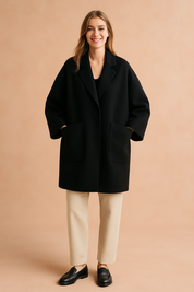 Oversized black wool coat with wide sleeves, large front pockets, and relaxed fit, paired with beige trousers and black loafers for a sleek minimalist style.