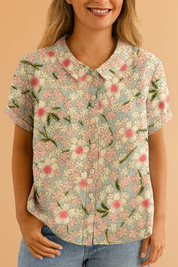 Short-sleeved blouse with a rounded collar and button front, featuring a pastel floral pattern with pink and white blossoms, green leaves, and a light blue background on soft-textured fabric.