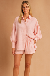A soft-textured pastel pink blouse and matching shorts set, designed with delicate woven patterns, button-up front, and voluminous sleeves. The coordinated outfit combines comfort and elegance, styled to highlight the breathable fabric's refined finish—perfect for relaxed yet chic daywear.
