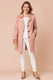 Light pink trench coat with button front, tailored fit, and side pockets, styled over a plain white top and paired with slim white trousers and taupe high heels.