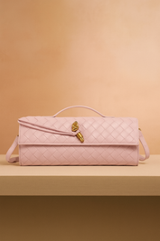 Rectangular pastel pink woven leather handbag with gold knot detail and flap closure, displayed on a beige platform against a warm blush gradient background.
