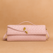 Pastel pink woven leather handbag with top handle, gold-tone hardware, and slim rectangular silhouette, placed on a warm sand and blush-toned gradient background.