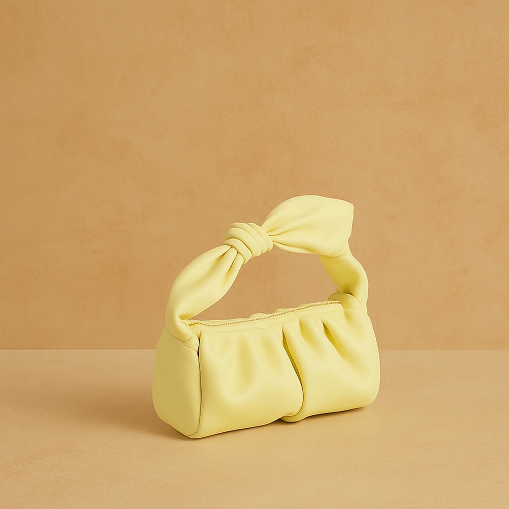A small pastel yellow handbag with a knotted handle, displayed on a warm sand surface with a soft blush gradient background in studio lighting.