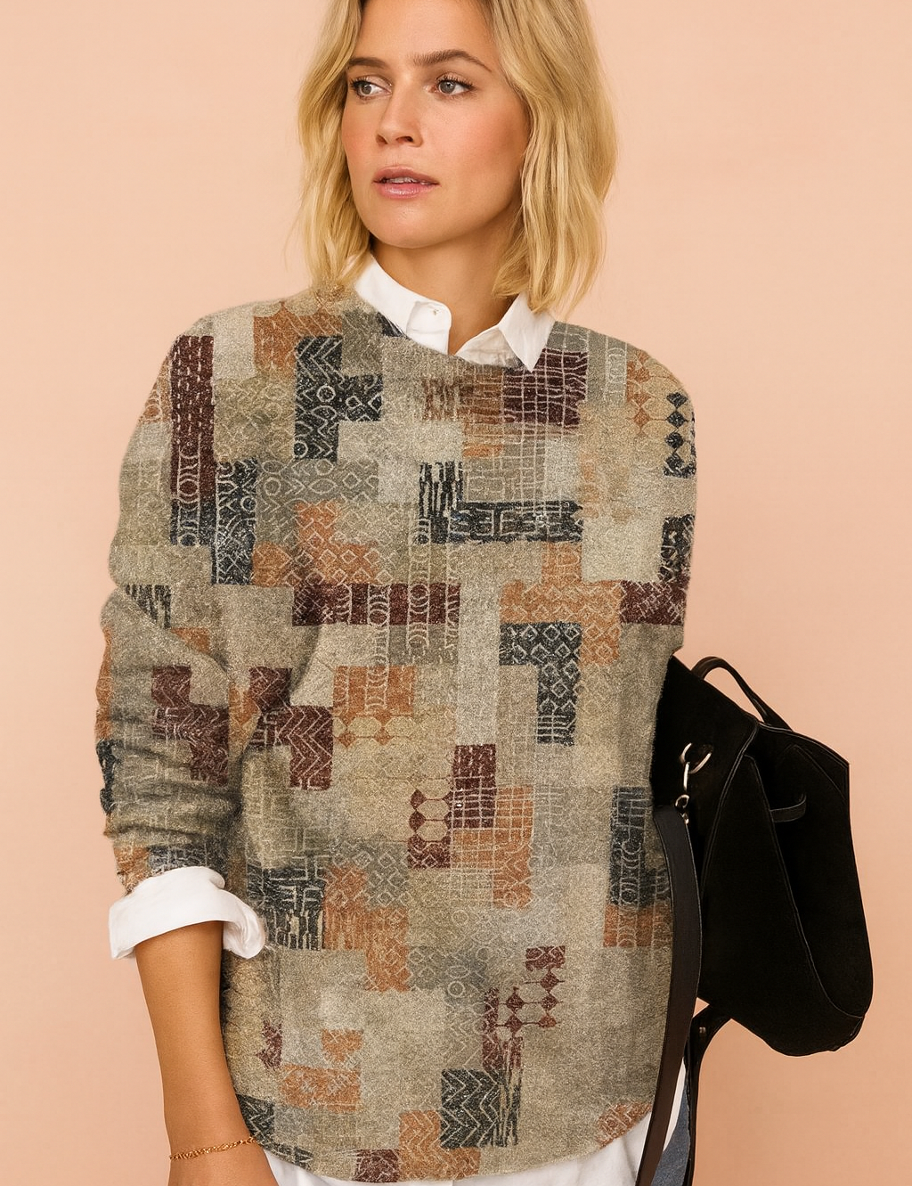 Patchwork pullover sweater with geometric patterns in muted tones of grey, rust, and navy, layered over a white collared shirt, paired with light blue jeans and a black suede handbag.