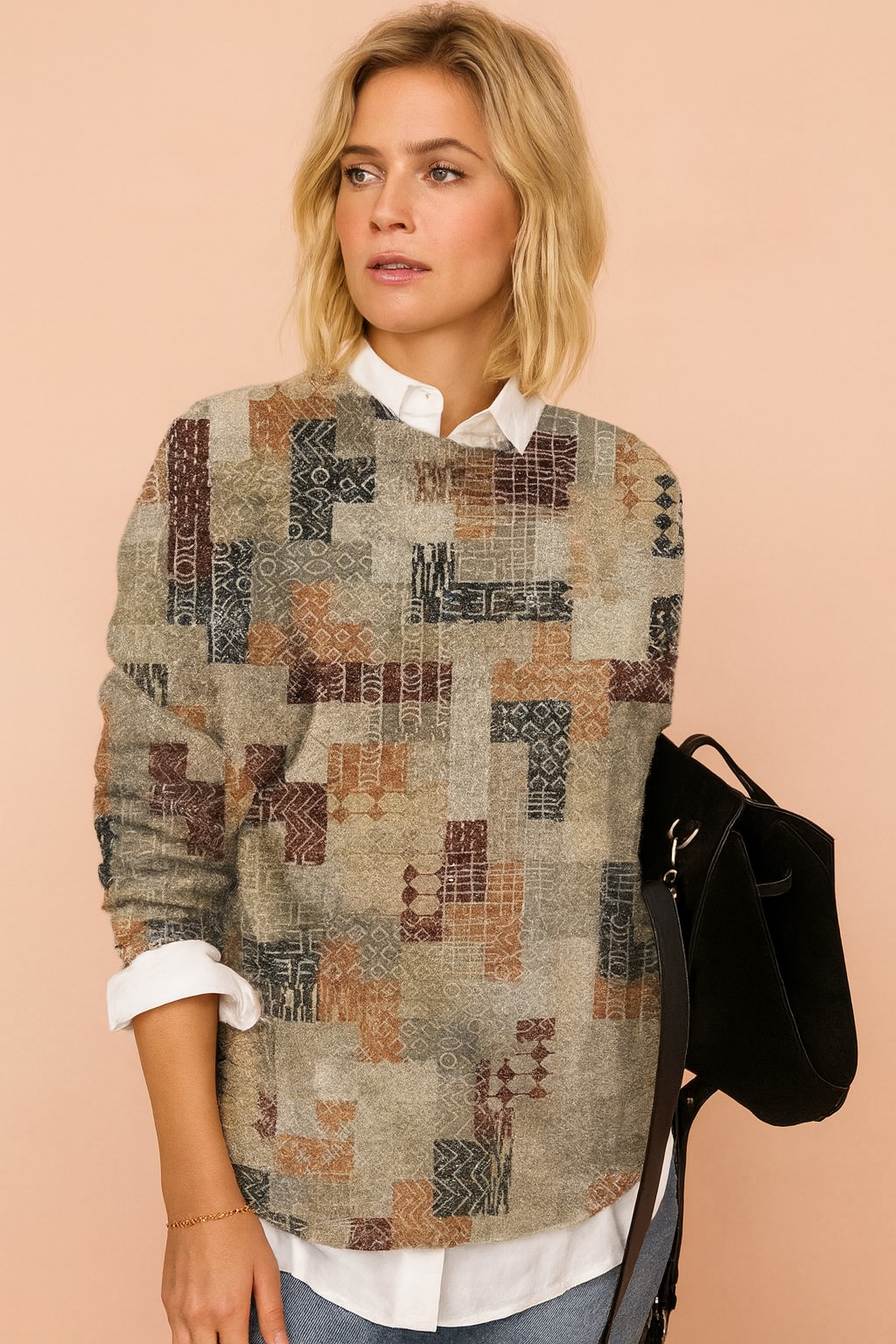 Patchwork pullover sweater with geometric patterns in muted tones of grey, rust, and navy, layered over a white collared shirt, paired with light blue jeans and a black suede handbag.