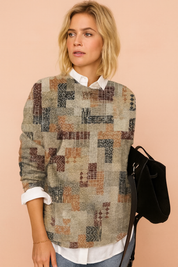 Patchwork pullover sweater with geometric patterns in muted tones of grey, rust, and navy, layered over a white collared shirt, paired with light blue jeans and a black suede handbag.