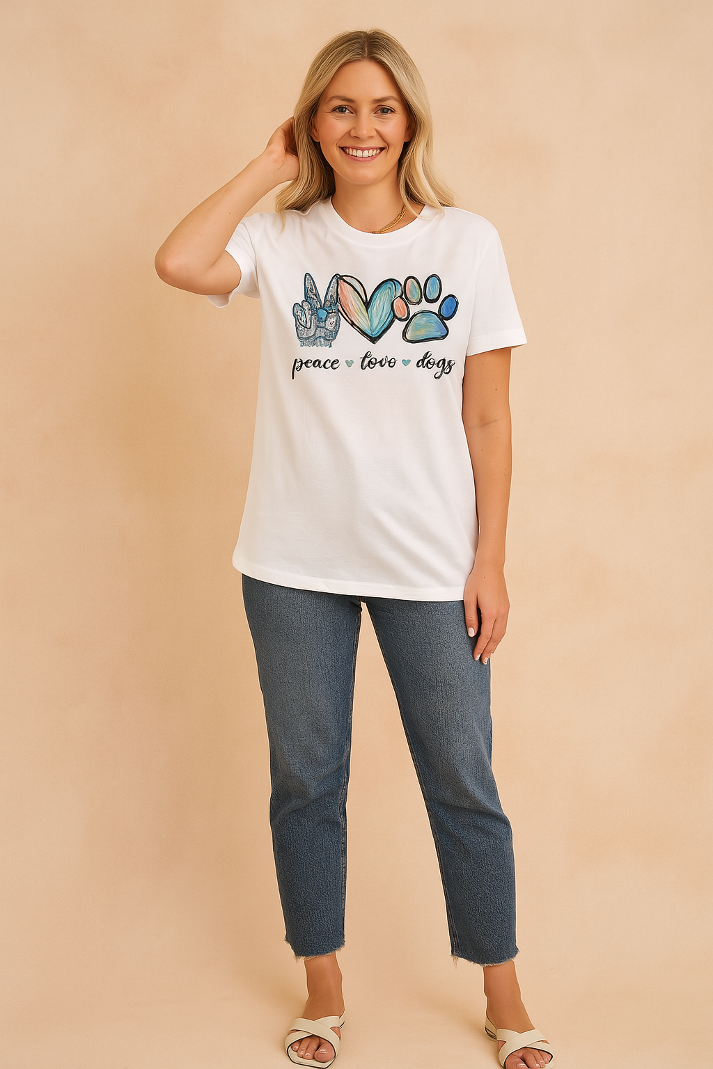 White graphic T-shirt with colorful "peace love dogs" print featuring a peace hand sign, heart, and paw print, styled with blue jeans and white crossover sandals.