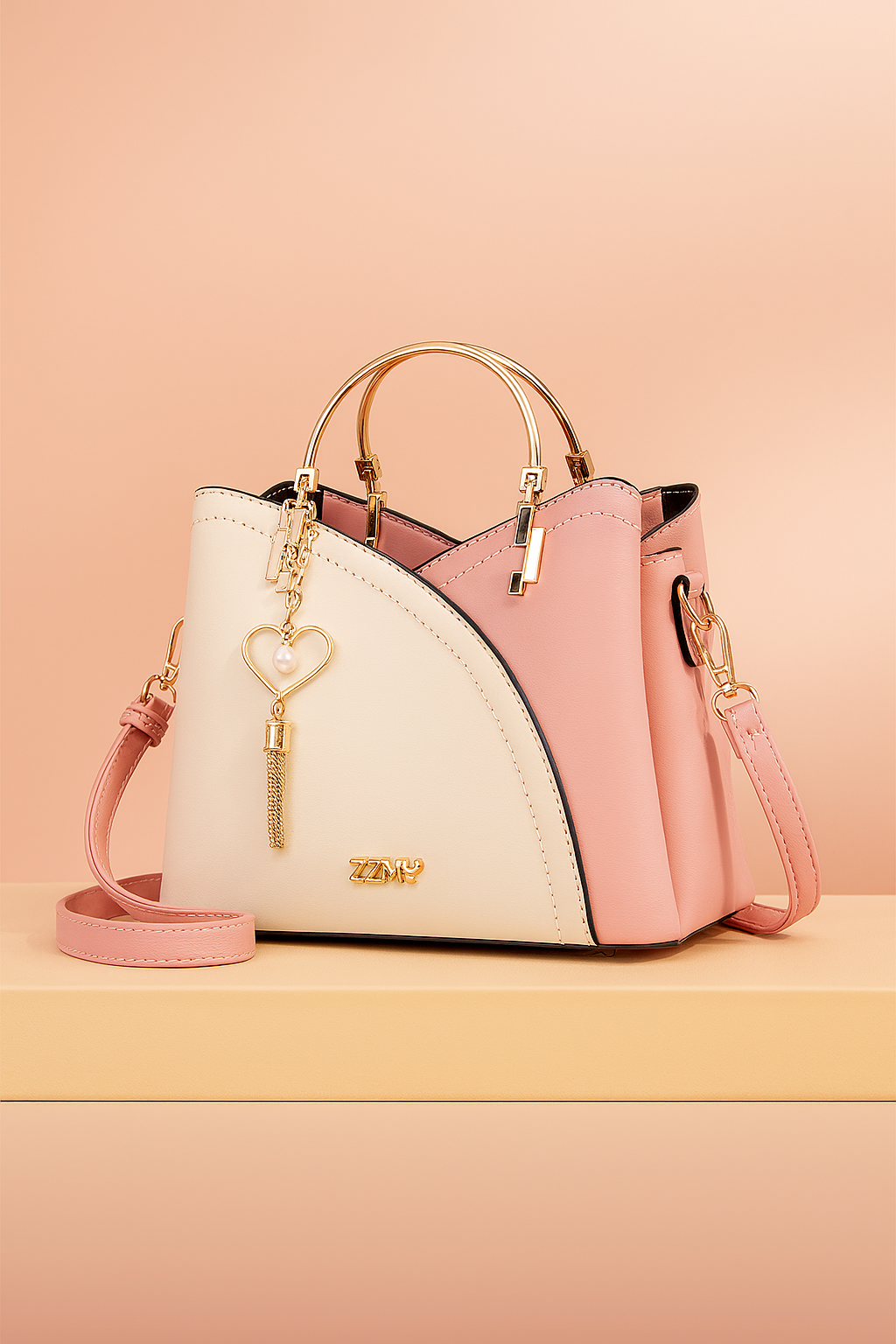 Two-tone pink and cream leather handbag with curved panel design, gold top handles, heart-shaped pearl tassel charm, and detachable strap, styled on a beige platform with a soft blush gradient background.