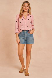 Pink linen button-up shirt featuring black swallow bird print and white buttons, styled with rolled-up sleeves and paired with high-waisted mid-wash denim shorts.