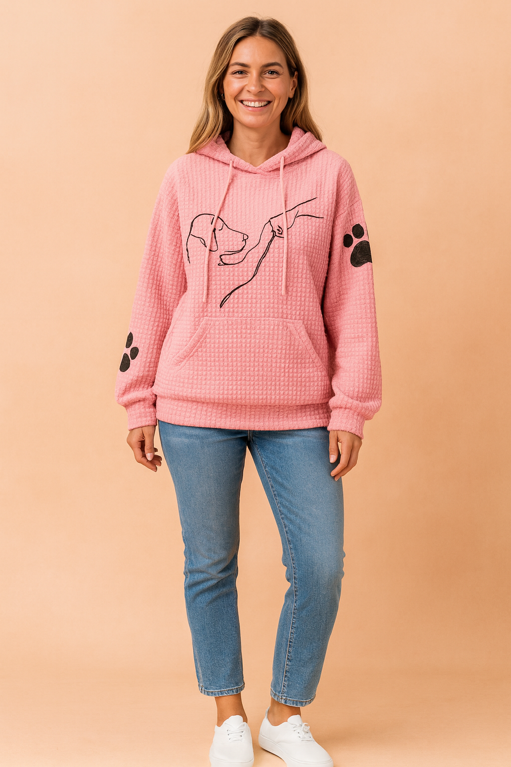 Textured pink waffle knit hoodie with minimalist black dog and human hand line art on the chest, black paw prints on each sleeve, kangaroo front pocket, and hood with drawstrings, styled with slim blue jeans and white sneakers.