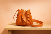 Pleated burnt orange bucket bag with gold hardware, drawstring-style top, and dual shoulder straps, made from textured faux leather and displayed on a light wood platform.