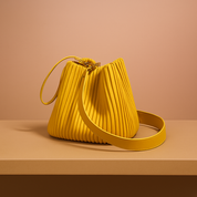 Mustard yellow pleated leather bucket bag with gold hardware, featuring a top wristlet and wide shoulder strap, positioned on a beige platform against a soft blush and sand gradient background.