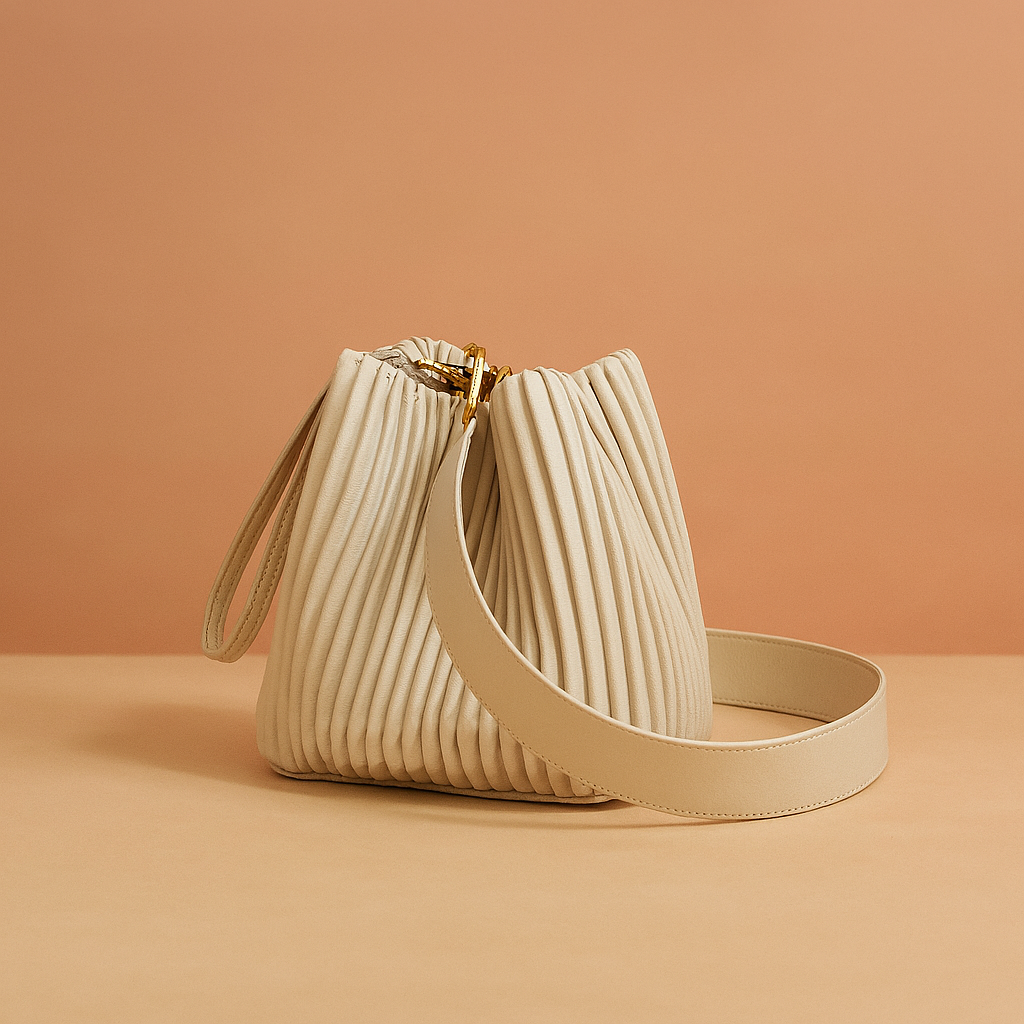 Luxurious off-white pleated leather bucket bag with gold hardware, top wristlet loop, and wide shoulder strap, displayed on a warm beige platform against a soft sand-to-pink gradient backdrop.