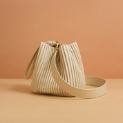 Luxurious off-white pleated leather bucket bag with gold hardware, top wristlet loop, and wide shoulder strap, displayed on a warm beige platform against a soft sand-to-pink gradient backdrop.