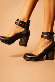Black leather block heel pumps with dual ankle straps and back zippers, featuring slightly worn edges and dark metal buckles for a vintage-inspired finish.