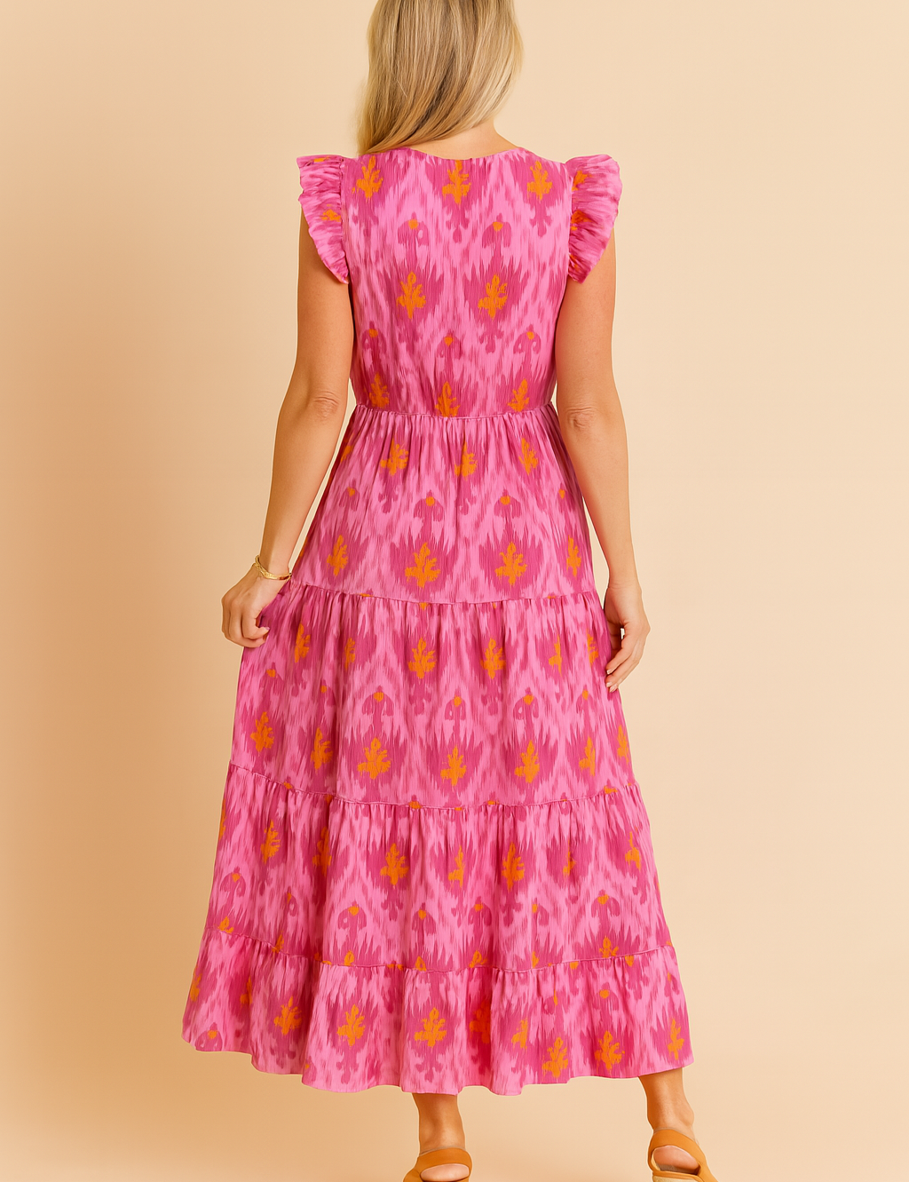 Back view of a tiered pink maxi dress featuring a vivid orange ikat-style flame motif, structured ruffle sleeves, a defined waistline, and soft flowing fabric.