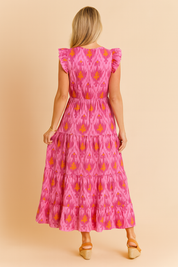 Back view of a tiered pink maxi dress featuring a vivid orange ikat-style flame motif, structured ruffle sleeves, a defined waistline, and soft flowing fabric.