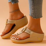Beige wedge thong sandals with perforated straps and large gold buckle accents, featuring a thick cushioned sole and subtle embossed side logo.
