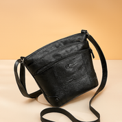 Black leather crossbody bag with front zipper pocket and subtle logo detail, placed on a warm surface with a softly lit sand and pastel pink gradient background.