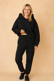 Black fleece coord set featuring a structured oversized hoodie with a deep front hood and dropped shoulders, paired with high-waisted joggers with a gathered elastic waistband and cuffed ankles. Both pieces are made from thick, matte cotton-blend fabric with a smooth, uniform texture and no visible prints or embellishments.
