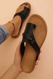 Minimalist black leather slip-on sandals with toe loop and cross-strap detailing, featuring fine stitching and a single metal rivet on a tan footbed.
