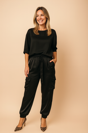 Two-piece black satin co-ord set featuring a relaxed-fit top with elbow-length sleeves and matching high-waisted jogger-style cargo pants with side pockets and a drawstring waist, paired with leopard print heels.

