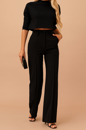 High-waisted black tailored trousers with a front crease, side pockets, and a matching black cropped top with elbow-length sleeves and a mock neck, styled with black stiletto sandals and a clutch.