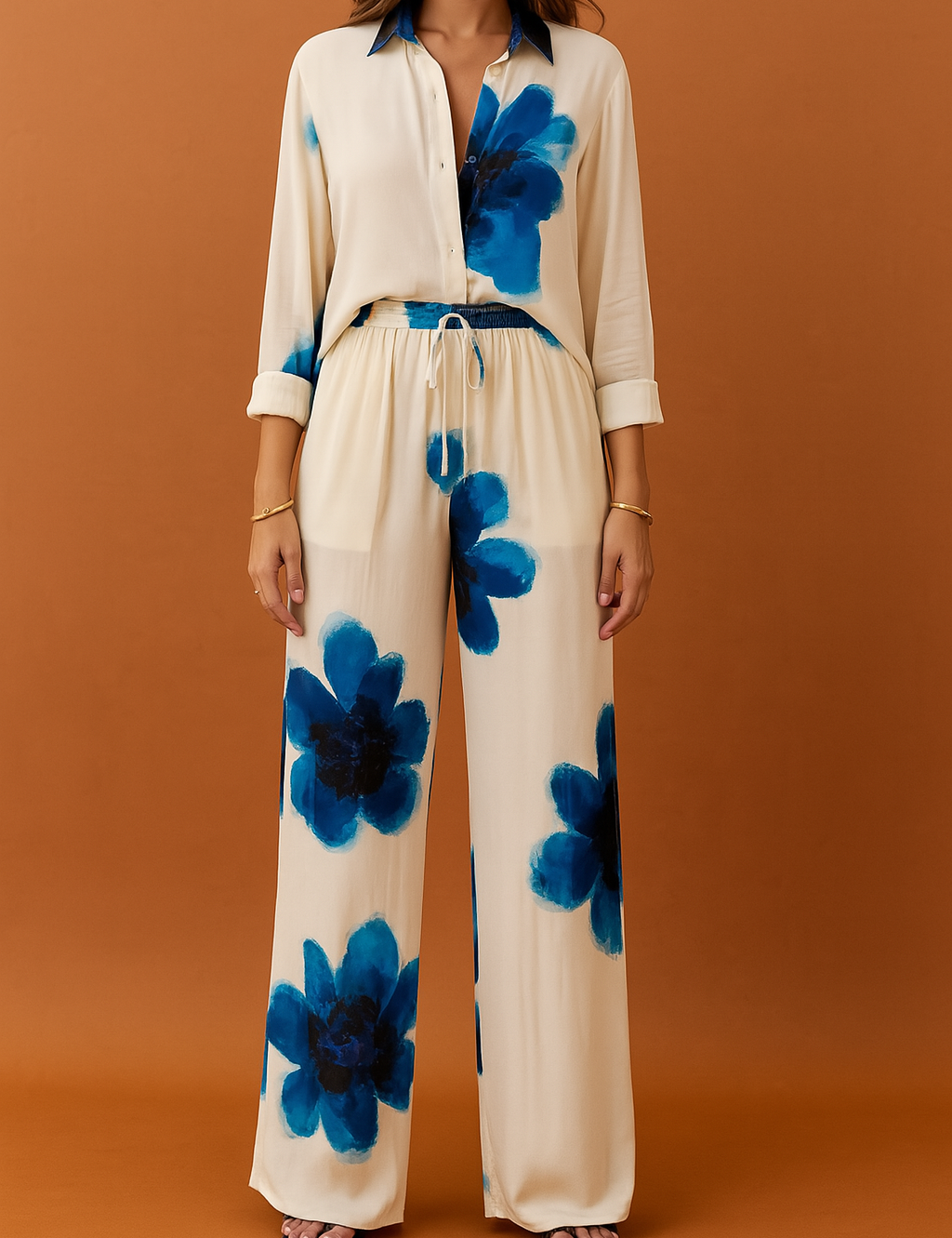 Cream two-piece coord set featuring a long-sleeve button-down shirt and wide-leg drawstring trousers, both adorned with oversized blue watercolor floral prints. Crafted from lightweight, fluid fabric with a soft drape and subtle sheen, offering a bold yet elegant daytime resort look.

