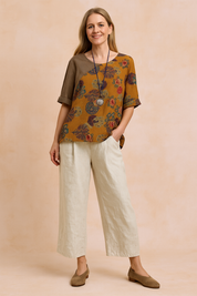 Loose-fit asymmetric blouse with rich amber-orange base, featuring colorful fan and floral prints in purple, blue, and red, button-accented at the shoulder and paired with neutral beige wide-leg trousers.