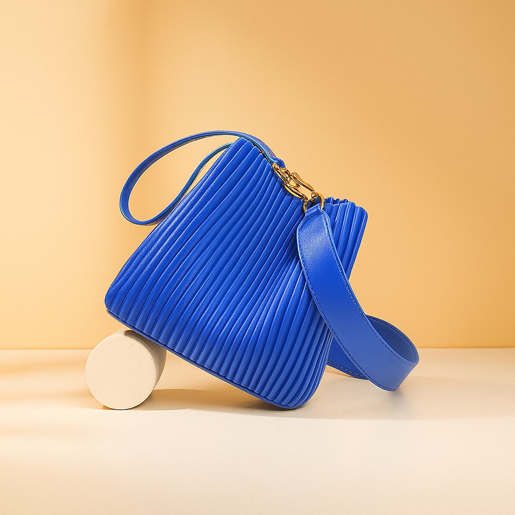 Cobalt blue pleated handbag with gold clasp and structured wide strap, displayed on a cream-toned surface with a soft gradient background in warm sand and pastel blush tones.