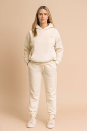 Cream fleece hoodie and matching sweatpants set featuring a relaxed fit, subtle embroidered chest detail, and ribbed cuffs and hems. The ensemble is completed with low-top cream sneakers for a coordinated casual look.