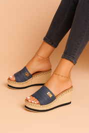 Denim slide wedge sandals with jute-wrapped platform heels, gold emblem accents, and a textured fabric upper in a rich navy hue.