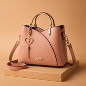 Blush pink faux leather handbag with gold-tone “ZZMY” logo, white contrast stitching, gold heart-shaped pearl tassel charm, and elegant gold hardware, styled on a warm beige platform in soft studio lighting.