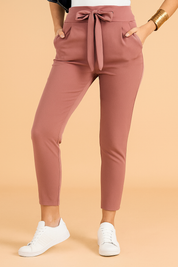 High-waisted dusty rose tapered pants with a wide waistband, self-tie belt detail, and slanted side pockets, crafted in a smooth, matte stretch fabric for a polished casual look.