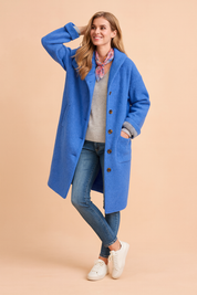 Bright blue button-up wool coat with rolled cuffs, side pockets, and long straight cut, styled over a light sweater and denim jeans.