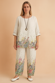 Relaxed-fit white linen loungewear set featuring a vibrant multicolor wildflower print at the hem of the top and bottom, with elbow-length sleeves and wide-leg pants, styled with a vintage pendant necklace.