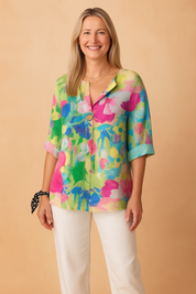 Loose-fit linen blouse featuring a vibrant watercolor floral print in shades of magenta, lime green, cobalt blue, and soft white, designed with a front button detail and elbow-length sleeves.