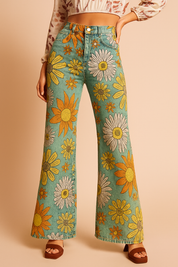 High-waisted wide-leg jeans with bold oversized daisy print in white, yellow, and orange on a faded teal base, styled with a boho crop blouse and brown open-toe heels.