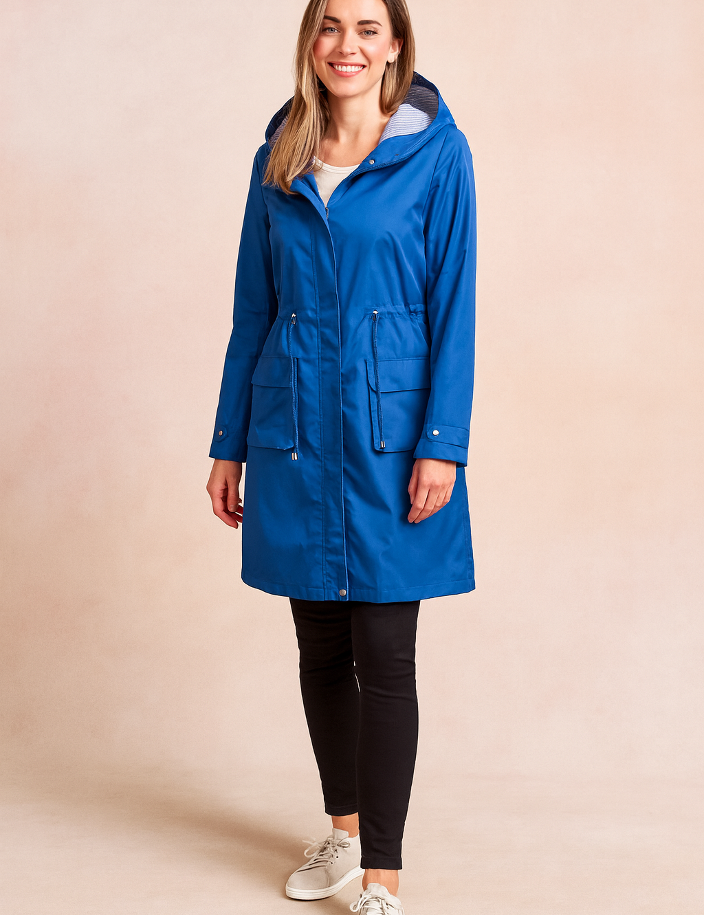 Women's bright cobalt blue long raincoat with front zip and snap button closure, adjustable waist drawcord, large front flap pockets, and striped hood lining, paired with black leggings and white sneakers.