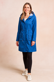 Women's bright cobalt blue long raincoat with front zip and snap button closure, adjustable waist drawcord, large front flap pockets, and striped hood lining, paired with black leggings and white sneakers.
