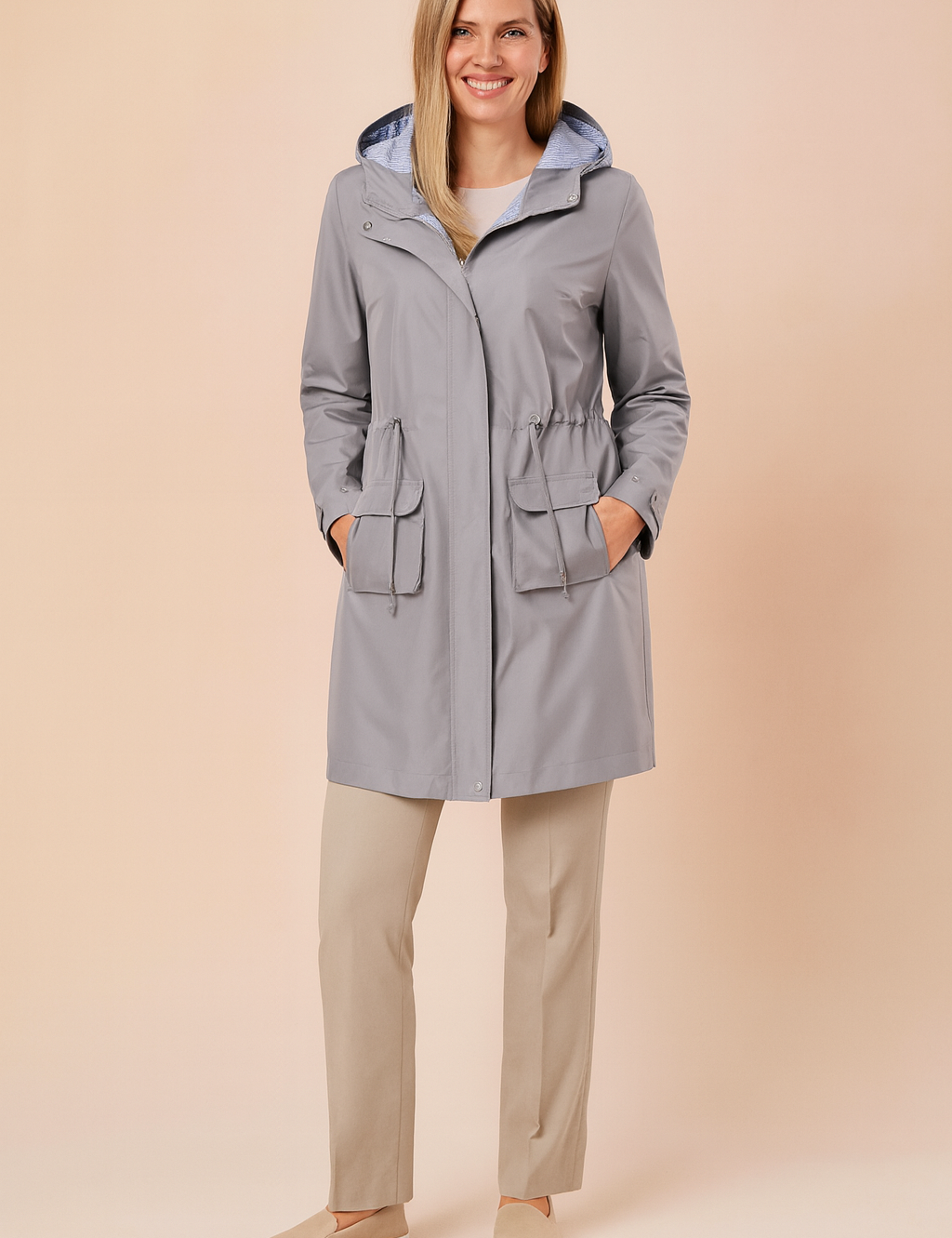 Light grey hooded raincoat with striped hood lining, front zipper, adjustable waist drawcord, and two large flap pockets, paired with straight-leg beige trousers and beige slip-on shoes.

