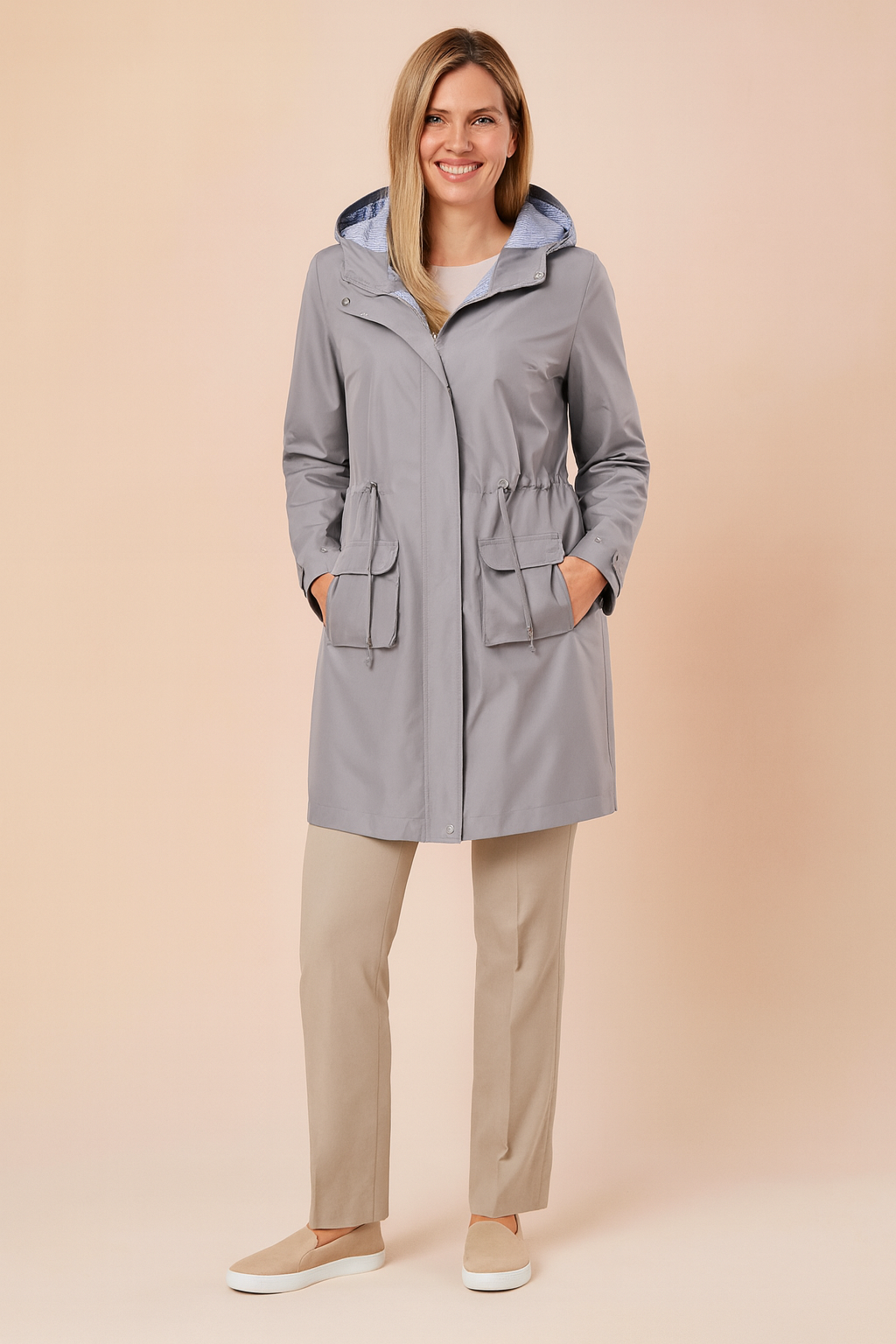 Light grey hooded raincoat with striped hood lining, front zipper, adjustable waist drawcord, and two large flap pockets, paired with straight-leg beige trousers and beige slip-on shoes.


