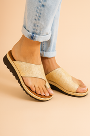 Gold woven slide sandals with crossover straps and black ridged soles, featuring a natural textured fabric and toe-loop detail.