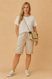 Light beige drawstring knee-length linen shorts paired with a loose white T-shirt, styled with white low-top sneakers and a straw crossbody bag with black trim.